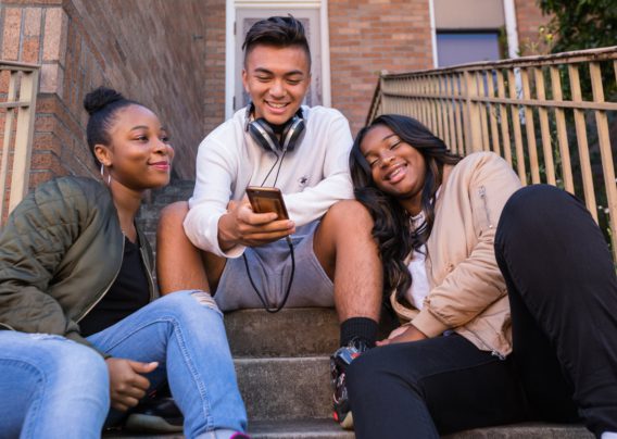 Three friends seated on an outdoor staircase, with a young man in the center holding a mobile phone and smiling at the screen. The two young women on either side are also looking at the phone screen