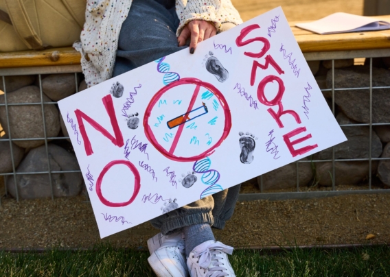 A person wearing jeans and a white cardigan holds an anti-smoking protest sign. Only the sign and their legs are visible in the photo, with their head out of frame.