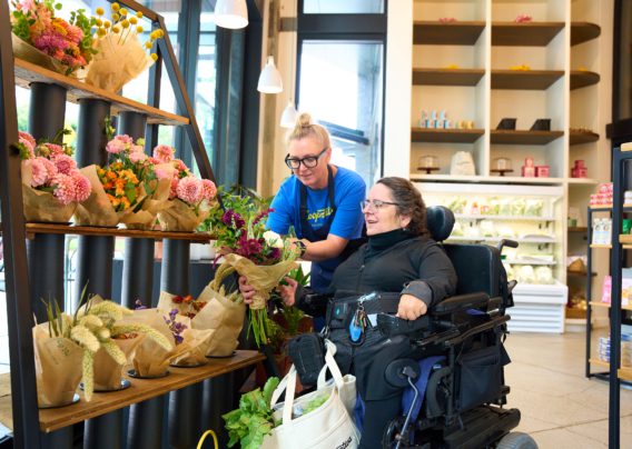A woman in a wheelchair, dressed in a black jacket and pants, examines flowers for sale at a small grocery store. She is assisted by a female employee wearing a blue apron, who bends down to touch the flowers the woman is considering.